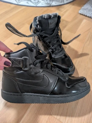 Nike - Svarta Nike sneakers med glansig finish och fuskpäls på plösen. Klassisk snörning och rem med spänne runt ankeln. Snygg, rund tå och platt sula. Perfekt för dig som gillar streetstyle och vill ha extra värme under vintern. Endast använd ett fåtal gånger. Som ny.