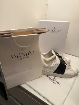 Valentino Garavani Open sneakers  - Säljer ett par Valentino Garavani Open sneakers i vitt skinn med bred svart läderdetalj över mitten. Skorna har snörning, rund tå och platt sula med nitar längs kanten. Kommer med originalkartong och dustbag. Perfekt för dig som gillar exklusiva sneakers.