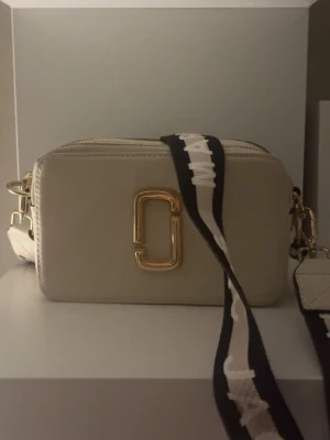 Beige axelväska från Marc Jacobs - Säljer en beige axelväska från Marc Jacobs i skinn med guldfärgad logodetalj framtill. Väskan har en bred, svart och beige axelrem med vit text, dubbla dragkedjor upptill och en praktisk ficka på baksidan. Perfekt storlek för det viktigaste och snygg design.