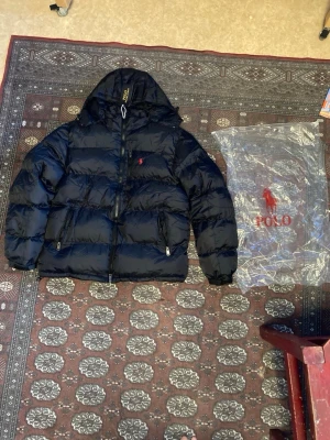 Svart pufferjacka från Polo Ralph Lauren - Säljer en svart pufferjacka från Polo Ralph Lauren med huva och dragkedja framtill. Jackan har två sidofickor med dragkedja och den klassiska röda Polo-loggan broderad på bröstet. Perfekt för kalla dagar och riktigt snygg till streetwear-stilen.
