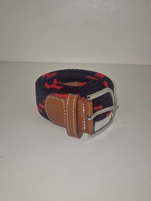 Svart och röd flätad bälte - Snyggt flätat bälte i svart med röda detaljer och brunt läder vid spännet. Bältet har ett klassiskt metallspänne och kontrastsömmar på läderdetaljerna. Perfekt för att ge outfiten lite edge och färg.