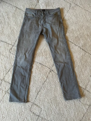 Grå skinny jeans från Hugo Boss - Säljer ett par gråa skinny jeans från Hugo Boss med klassisk femficksdesign och knappgylf. Jeansen har jätte snygg passform och är tillverkade i mjukt bomullsmaterial med lite stretch för extra komfort. Perfekt för dig som gillar en stilren och enkel look.