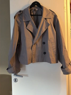 Grå croppad trenchcoat från H&M Divided - Snygg grå croppad trenchcoat från H&M Divided med bred krage, dubbelknäppning och detaljerade knappar på axlar och ärmslut. Jackan har lång ärm och är gjord i bomullsmaterial. Perfekt för dig som gillar trendiga och klassiska plagg.