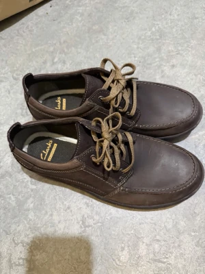 Bruna seglarskor från Clarks, EU 42 - Säljer ett par bruna seglarskor från Clarks i storlek 42. Skorna har snörning, rund tå och är tillverkade i skinn med kontrastsömmar. Perfekta för dig som gillar klassisk stil och vill ha något som funkar till jeans eller chinos.
