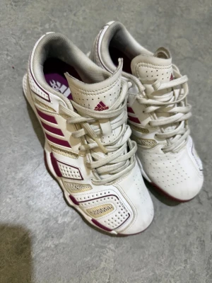 Adidas Adizero vita sneakers med lila - Säljer ett par Adidas Adizero sneakers i vitt med lila detaljer och klassiska ränder på sidorna. Skorna har snörning, perforerad ovandel och lila innersula med Adidas-logga. Materialet är syntet och skinn, perfekt för dig som gillar sportig stil.