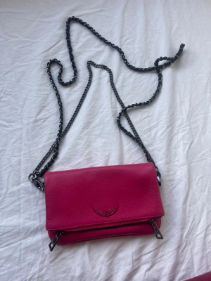 Rosa axelväska med kedja från Zadig & Voltaire.  - Säljer en snygg rosa axelväska från Zadig & Voltaire i skinn med silverfärgad kedja och dragkedja. Väskan har ett emblem med vingar framtill och flera fack med dragkedja. Perfekt för dig som vill ha en cool och färgstark accessoar. Både kort och långt band. Dustbag kommer till och den är nästintill oanvänd