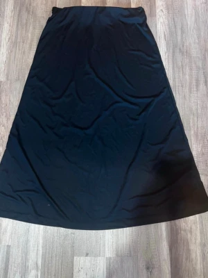 Svart långkjol i mjukt tyg - Säljer en svart långkjol med enkel och stilren design. Kjolen har en A-linjeformad siluett och är gjord i ett mjukt, följsamt material som känns skönt mot huden. Perfekt att matcha med sneakers eller boots för en chill look.