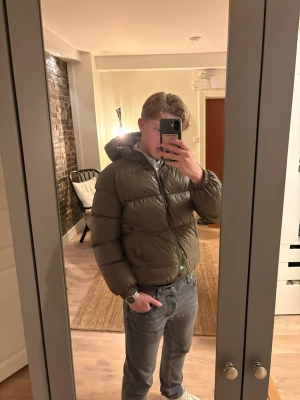 Olivgrön pufferjacka med huva - Säljer en sprillans ny olivgrön pufferjacka med huva och dragkedja framtill. Jackan har en quiltad look med breda kanaler och elastiska muddar vid ärmsluten. Perfekt för kyliga dagar och riktigt skön att bära. Storlek: S passar XS