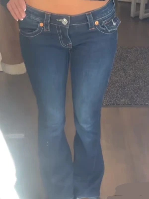 Mörkblå True Religion jeans bootcut - Säljer ett par mörkblå jeans från True Religion med bootcut-snitt och låg midja. Jeansen har tydliga vita kontrastsömmar, stora fickor med lock och silverfärgade knappar. Klassisk True Religion-logga på bakfickan och patch i midjan. Perfekt för dig som gillar detaljer.