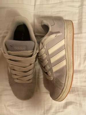 Ljusgrå adidas sneakers i mocka - Säljer ett par ljusgrå adidas sneakers med klassiska vita ränder på sidorna och vit sula med gummiyta undertill. Skorna har snörning och är tillverkade i mjuk mocka. Perfekta för dig som gillar stilrena och bekväma skor med retrovibe.