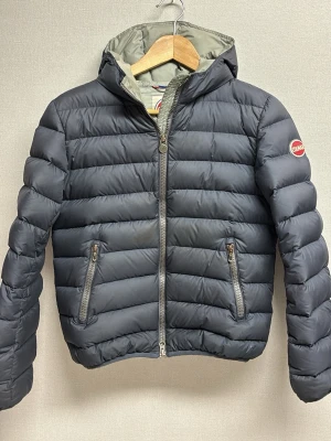 Mörkblå Colmar vinterjacka - Säljer en mörkblå pufferjacka från Colmar med huva och två sidofickor med dragkedja. Nackdelen med denna jacka är att det står namn och telefonnummer till tidigare ägare som man ser på den fjärde bilden. Detta är förstås en sak som sänker priset men egentligen så gör det inte så jättemycket eftersom jackan annars är som den ska, bara någon fläck på insidan men annars är jackan i bra skick.