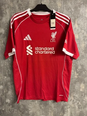 Liverpool Isak - Ny storlek XL (liten fläck se bild) - Bilderna visar en röd Liverpool fotbollströja med vita detaljer, Adidas-logga och tryck 'ISAK 9' på ryggen. Ingen sko finns med på bilderna. Liten svart fläck se bild