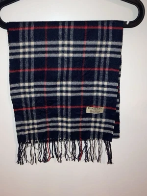 Rutig ullhalsduk från Burberry - Säljer en klassisk rutig halsduk från Burberry i 100% lammull. Färgerna är mörkblå, vit och röd med fransar i ändarna. Perfekt för detta kalla väder.