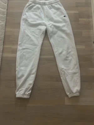 Vita mjukisbyxor från Champion - Säljer ett par vita mjukisbyxor från Champion med elastisk midja och muddar vid bensluten. Byxorna har sidofickor och en bakficka, samt den klassiska Champion-loggan broderad på ena sidan. Perfekta för chill dagar eller när du vill vara extra bekväm.