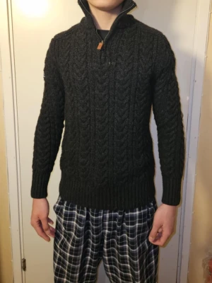Mörkgrå stickad Superdry tröja half zip - Säljer en mörkgrå stickad tröja från Superdry med grovt kabelmönster och hög krage med half zip. Tröjan har långärmade ribbade muddar och en liten Superdry-patch på ärmen. Perfekt för kyliga dagar och riktigt snygg till jeans.