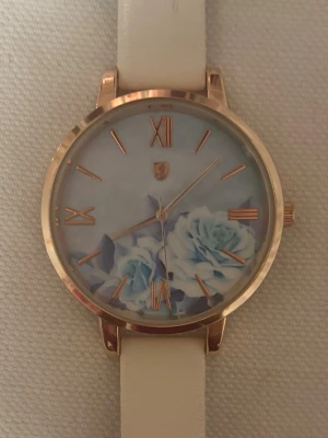 Blommig klocka med vit rem - Snygg klocka med blommigt motiv på urtavlan i blå och vita toner. Urtavlan har roséguldfärgade romerska siffror och detaljer. Remmen är vit. Perfekt accessoar för att lyfta din outfit. Klockan behöver byttas batteri för att fungera.