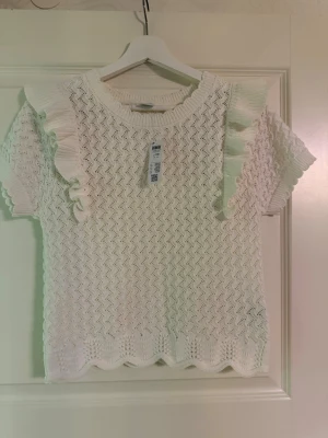 Vit stickad topp med volang från Lindex - Supersöt vit stickad topp från Lindex i storlek 170. Den har korta ärmar, volangdetaljer på axlarna. Storleken kan även passa till xs
