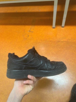 Svarta Nike Air Force 1 sneakers - Säljer ett par klassiska svarta Nike Air Force 1 sneakers med låg profil. Skorna har snörning, rund tå och platt sula. Tillverkade i skinn med diskret Nike-logga på sidan. Perfekt för dig som gillar stilrena och tidlösa sneakers.