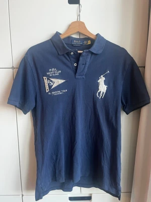Marinblå pikétröja från Polo Ralph Lauren - Säljer en marinblå pikétröja från Polo Ralph Lauren, custom slim fit. Tröjan har korta ärmar, krage och knappar framtill. På bröstet finns broderade detaljer med logga och text, samt en vit polospelare. Perfekt för en sportig och stilren look. Hör av er om frågor. 