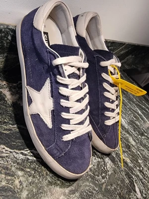 Golden Goose Super-Star blå sneakers - stl 40 - Golden Goose Super-Star sneakers i mörkblå mocka med vit stjärndetalj på sidan och vita skinnpartier vid häl och snörning. Klassisk rund tå och platt sula och vita snören. Stl. 40. Vissa tycker de är stora i storleken. Se Golden Goose hemsida för storlekstabell. Välbehållet skick, normalt slitag lite skavda under häl - se bild) Originalkartong, dustbag samt äkthetsbevis ingår. 