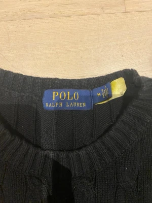 Svart stickad tröja Polo Ralph Lauren - Svart stickad tröja från Polo Ralph Lauren i storlek M. Klassisk kabelstickad design med rund hals och långa ärmar. Tröjan har en liten röd broderad logga på bröstet och ribbade muddar vid ärmslut och nederkant. Perfekt för dig som gillar stilrena och tidlösa plagg.