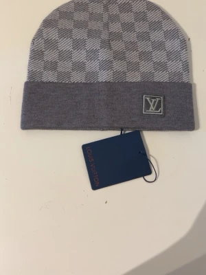 Grå rutig lv väst - Säljer en grå väst från Louis Vuitton med klassiskt rutigt mönster och LV-logga framtill. Västen är stickad och har en exklusiv känsla med snygga detaljer. Perfekt för dig som gillar designerplagg och vill sticka ut med stil.