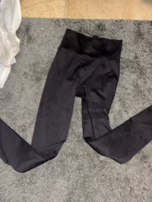 Svarta leggings från Stronger - Säljer ett par svarta leggings från Stronger med ribbad struktur och bred midja. De har diskreta ränder och är tillverkade i ett stretchigt syntetmaterial som sitter tight och bekvämt. Perfekta för träning eller chill dagar.