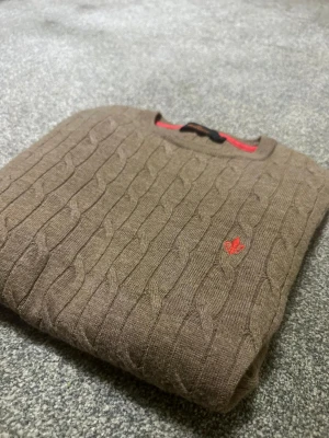 Brun kabelstickad tröja Morris - Brun kabelstickad tröja från Morris med rund hals och snygga röda detaljer, bland annat ett broderat emblem på bröstet och röda armbågslappar. Tröjan har en klassisk look och är perfekt för dig som gillar stilrena plagg med lite edge.