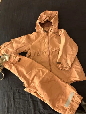 Skal överkläder från Newbie - Säljer ett par persikofärgade täckbyxor från Newbie i storlek 98. Byxorna har justerbara hängslen, resår i midjan och reflexdetaljer vid benslut. Materialet är slitstarkt och passar perfekt för utomhusaktiviteter under kyligare säsonger.