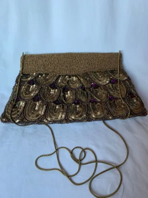 Glittrig aftonväska från Oscar & Clothilde - Säljer en paljettbeströdd kuvertväska från Oscar & Clothilde med pärlbroderad ovandel och paljettmönster i guld och lila detaljer. Väskan har ett långt guldfärgat axelband/metallkedja (kan stoppas in i väskan om man föredrar att ha den som kuvertväska) och magnetlås. Lite känsla av art nouveau eller art deco. Aldrig använd så i absolut toppskick. 20-25 cm bred, 14 cm hög. Kedjan är 114 cm lång. 