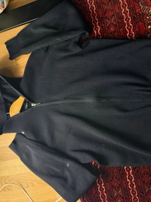 Mörkblå stickad hoodie från Qingwai - Säljer en mörkblå stickad hoodie från Qingwai med dragkedja framtill och huva. Tröjan har långärmade ribbade muddar och är i ett grovstickat material som känns både chill och stilrent. Perfekt för dig som gillar en avslappnad look med lite edge.