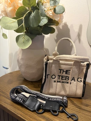 Beige tote bag från Marc Jacobs - Säljer en beige tote bag från Marc Jacobs ( EJ ÄKTA )med svarta detaljer och tydlig logga framtill. Väskan är i kraftig bomull och har både handtag och en avtagbar, randig axelrem med text. Perfekt storlek för det viktigaste 