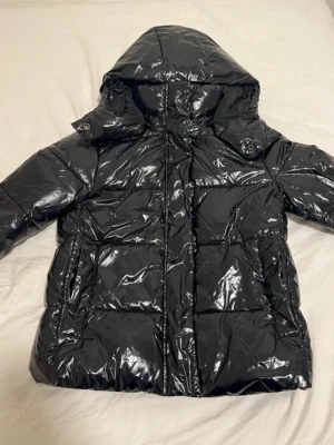 Svart pufferjacka från Calvin Klein - Säljer en svart, glansig pufferjacka från Calvin Klein Jeans med huva och dragkedja. Jackan har quiltad design och logga på bröstet. Perfekt för kalla dagar. Den är i väldigt bra skick då jag bara har använt den fåtal gånger❤️