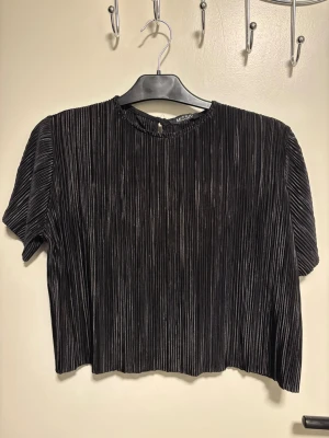 Svart plisserad croppad topp från Missa - Säljer en svart croppad topp från Missa med plisserad struktur och korta ärmar. Toppen har rund halsringning och en liten knapp i nacken. Materialet är glansigt och ger en cool vibe till outfiten.