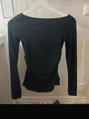 Svart långärmad topp från Bershka - Svart långärmad topp från Bershka med smal passform och lätt rynkning i midjan. Toppens material är mjukt och stretchigt, vilket gör den bekväm att bära. Enkel och stilren design med rundad hals och raka ärmar.