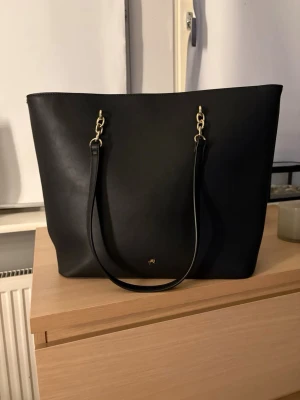 Svart axelväska från Anna Field - Fin Anna Field med guldfärgade kedjedetaljer på axelremmarna. Väskan är rymlig och har magnetknäppning samt ett litet innerfack med dragkedja. Den är som ny och är använd måste få gånger. 