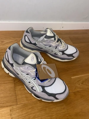Asics NYC-GEL Oister Gray Helt Nya!!! - Säljer ett par Asics GEL sneakers med snygg design i grått, vitt och ljuslila. Skorna har meshpaneler för bra ventilation, detaljer i syntet och skinn, samt en robust och dämpad sula. Klassisk snörning och rund tå. Perfekta för dig som gillar sportig stil.