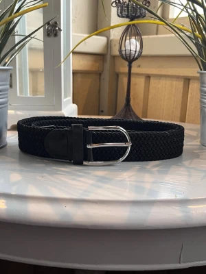 Svart flätat bälte - Bilderna visar ett svart flätat bälte med silverfärgat spänne, inte en väst. Ingen information om väst kan ges.