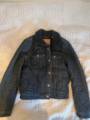 Svart jeansjacka med teddyfodring från Levi's - Säljer en svart jeansjacka från Levi's i storlek S med mjuk teddyfodring. Jackan har klassisk knappstängning framtill, två bröstfickor med lock och sidofickor. Perfekt för dig som gillar street och vill ha en cool look.