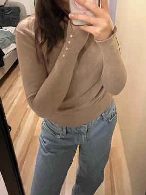 Beige långärmad stickad tröja - Snygg beige stickad tröja med lång ärm och ribbade muddar. Tröjan har dekorativa knappar vid ärmslutet som ger en extra detalj. Passformen är normal och materialet känns mjukt och bekvämt mot huden. Perfekt att matcha med jeans för en chill look.