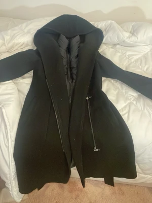 Svart kappa från Hollies med huva - Säljer en svart kappa från Hollies med stor huva och quiltad innerdel med dragkedja. Jackan har en stilren look och är gjord i ull och syntet, perfekt för kalla dagar. Märkespatch på ärmen och snygga detaljer med dubbla dragkedjor framtill.