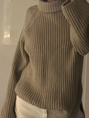 Beige ribbad polotröja stickad - Säljer en beige stickad polotröja med bred ribbad struktur. Tröjan har lång ärm, hög polokrage och en loose passform.