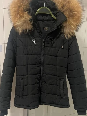 Svart pufferjacka med päls från Belle - Svart pufferjacka från Belle i storlek XS med fluffig päls på huvan. Jackan har quiltad design, dragkedja framtill och två sidofickor. Snygga detaljer med extra dragkedjor vid bröstet och justerbara snören på huvan. Perfekt för kalla dagar.