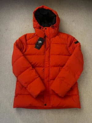 Röd dunjacka från J.Lindeberg, stl L - Säljer en fet röd dunjacka från J.Lindeberg i storlek L. Jackan har huva med snörning, dragkedja och knappar framtill samt quiltad design. Perfekt för kalla dagar och har en clean look med svarta detaljer på insidan av huvan. Originella pris 2800kr som ni kan se på bilden men kommer sälja den för 1053kr