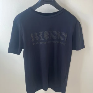 Svart BOSS t-shirt i bomull - Svart t-shirt från BOSS med diskret logga framtill i prickigt mönster. Klassisk rund hals och korta ärmar. Tillverkad i mjuk bomull för en skön känsla. Perfekt för dig som gillar stilrena och enkla plagg med en touch av premium.