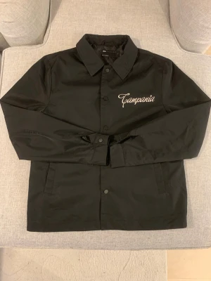 Svart overshirt från ASOS DESIGN - Svart overshirt från ASOS DESIGN med vit broderad text 'Campania' på bröstet och ryggen. Jackan har klassisk krage, knappar framtill och två sidofickor. Materialet är polyester och passformen är normal. Perfekt för dig som gillar stilrena och cleana plagg.