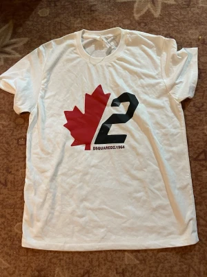 Vit DSQUARED2 t-shirt med tryck - Säljer en vit t-shirt från DSQUARED2 med stort tryck på bröstet som visar ett rött lönnlöv och siffran 2 i svart. Rund hals och korta ärmar, tillverkad i mjuk bomull. Perfekt för dig som gillar streetwear och snygga logotyper.