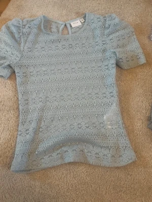 Ljusblå spetsblus från Vila - Säljer en ljusblå blus från Vila med korta puffärmar och rund hals. Blusen har ett vackert spetsmönster med blommiga detaljer och en liten knapp i nacken. Perfekt för dig som gillar romantisk stil och fina detaljer.