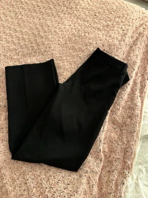 Svarta kostymbyxor med rak passform och lågmidjade - Snygga svarta kostymbyxor med klassisk rak siluett och midja med hällor för bälte. Materialet känns slätt och stilrent, perfekt för en clean look. Passar dig som gillar enkel och tidlös stil. Byxorna har en normal passning och är helsvarta utan mönster.