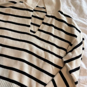 Randig stickad tröja från Gina Tricot - Beige och svart randig stickad tröja från Gina Tricot med v-ringning och krage. Tröjan har långa ärmar och en avslappnad passform, perfekt för lager-på-lager. Klassisk och tidlös design som funkar året runt.